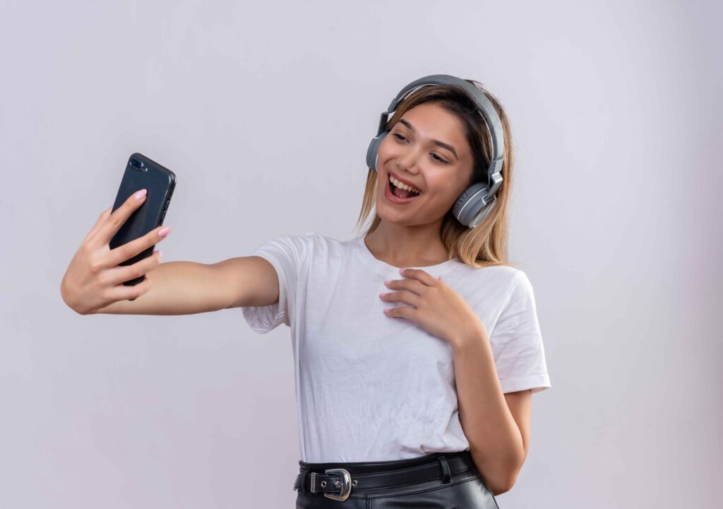 La imagen actual no tiene texto alternativo. El nombre del archivo es: una-mujer-joven-feliz-en-camiseta-blanca-usando-audifonos-tomando-un-selfie-con-smartphone-en-una-pared-blanca_11zon-scaled.jpg